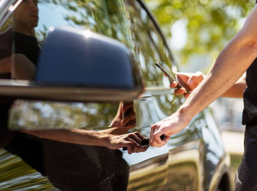 An auto locksmith opening a car using a keyless entry system while holding a smartphone to assist the process